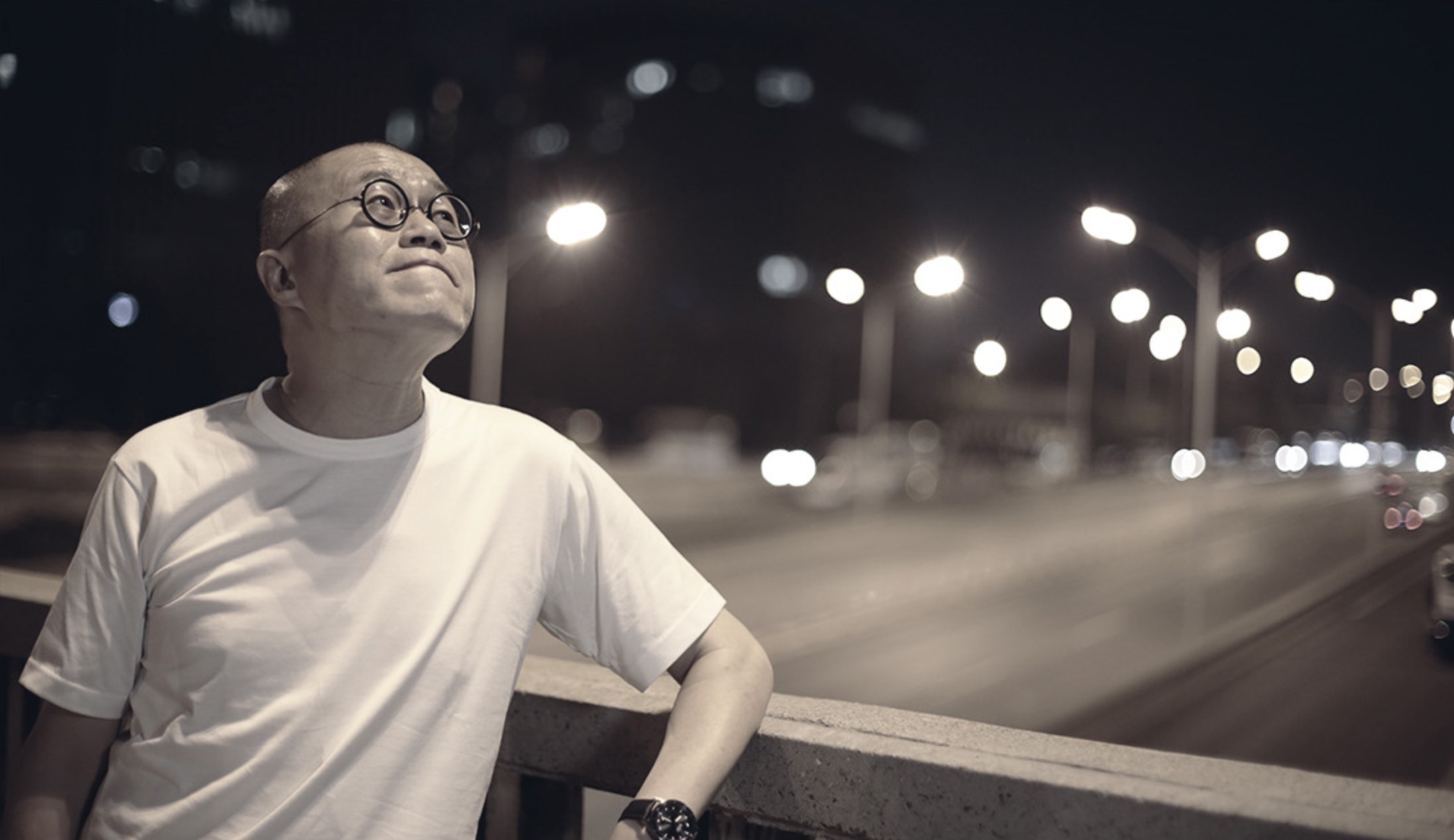 梁文道在夜晚街头阅读,身后有疾驰车辆和路灯
